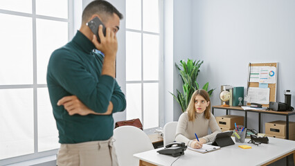 In an office, a man on a phone call stands while a focused woman writes at her desk, embodying workplace collaboration.
