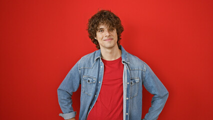 Handsome young man with curly hair smiling against a vibrant red background wearing a denim jacket.