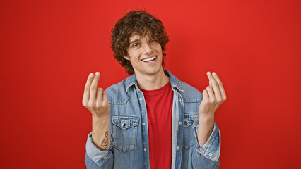 Young hispanic man with curly hair, smiling and making a gesture as if holding money, against a red...