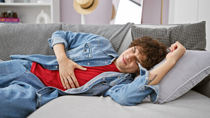 Hispanic man with curly hair, wearing denim shirt, feeling stomach pain on a grey sofa at home.