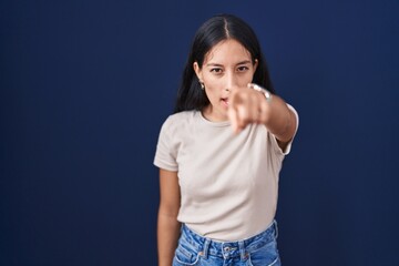 Young hispanic woman standing over blue background pointing displeased and frustrated to the...
