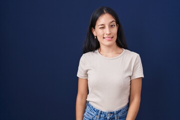 Young hispanic woman standing over blue background winking looking at the camera with sexy...