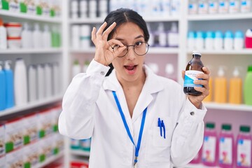 Young hispanic woman working at pharmacy drugstore holding syrup smiling happy doing ok sign with hand on eye looking through fingers