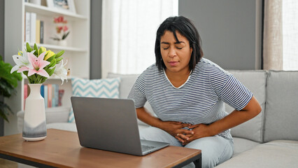 A middle-aged hispanic woman feeling abdominal pain while working on a laptop in a home living room.