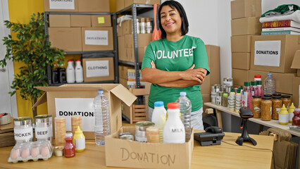 Smiling middle-aged hispanic woman volunteer standing in a donation center with food and clothes boxes.