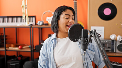 A hispanic woman singing joyfully in an orange music studio surrounded by instruments and recording equipment.