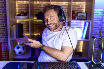 Middle age man with beard playing video games wearing headphones inviting to enter smiling natural with open hand