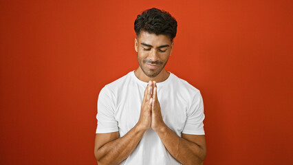 A young hispanic man with a beard in a white t-shirt against a solid red background expresses...