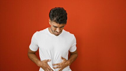 A young hispanic man grimaces in pain, clutching his stomach, isolated against a vibrant red wall.