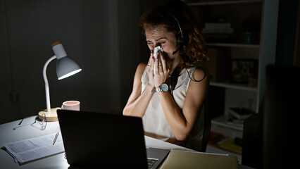 A stressed young woman with headphones at her night office desk wiping tears.