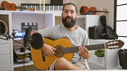Smiling man playing acoustic guitar in a home music studio surrounded by instruments and recording...
