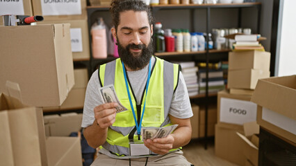 A bearded hispanic man in a vest counts money in an organized warehouse environment full of boxes.