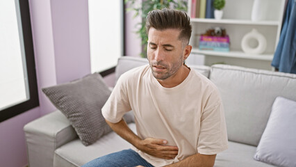 Obraz premium Worried young hispanic man suffering stomachache, touching aching belly while sitting on living room sofa. unhappy adult indoors, serious illness worry at home.