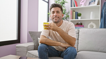 Confident young hispanic man enjoying his morning espresso, sitting comfortably on his living room sofa, a radiant smile blooming over his mug