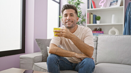 Fototapeta premium Confident young hispanic man enjoying his morning espresso, sitting comfortably on his living room sofa, a radiant smile blooming over his mug