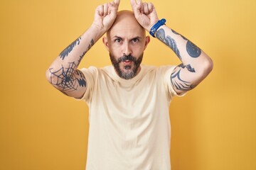 Hispanic man with tattoos standing over yellow background doing funny gesture with finger over head...