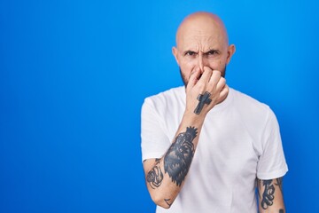 Hispanic man with tattoos standing over blue background smelling something stinky and disgusting, intolerable smell, holding breath with fingers on nose. bad smell