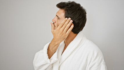 Handsome hispanic man in white bathrobe touching his face against an isolated light background.