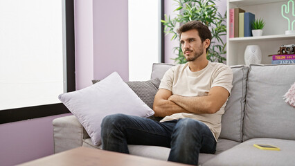Fototapeta premium A contemplative young hispanic man with a beard indoors, sitting on a grey sofa in a cozy home environment.