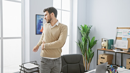 Naklejka premium Handsome hispanic man standing thoughtfully in a modern office setting, radiating professionalism and confidence.
