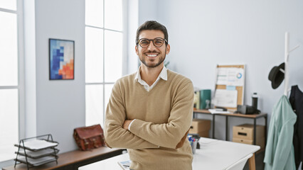Handsome young man with arms crossed standing confidently in a well-lit modern office interior.