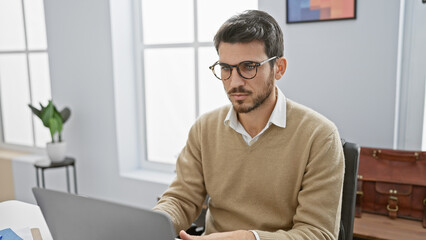 Handsome hispanic man with beard working at laptop in bright office room.