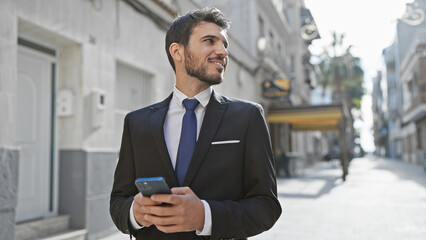Fototapeta premium Handsome young hispanic man in suit using smartphone on sunny urban street.