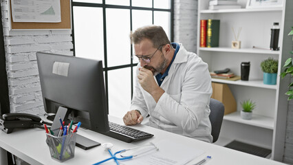 Mature man in lab coat coughing at desk with computer in clinic office.