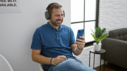 A handsome, mature hispanic man with grey hair enjoying music on headphones and smartphone in a modern room.