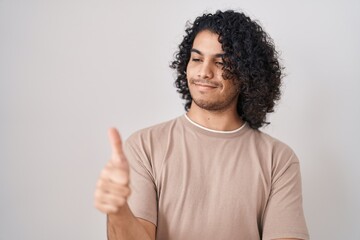 Hispanic man with curly hair standing over white background looking proud, smiling doing thumbs up...