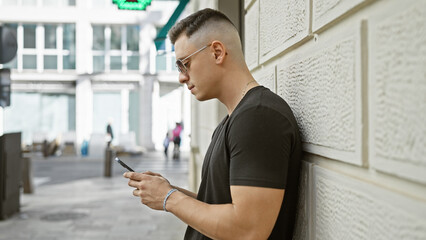 Handsome young hispanic man using smartphone on city street with blurred buildings background.