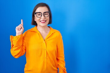 Middle age hispanic woman wearing glasses standing over blue background showing and pointing up with finger number one while smiling confident and happy.