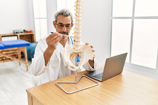 Middle age grey-haired man doctor holding anatomical model of spinal column at rehab clinic