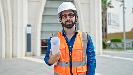 Young hispanic man architect smiling confident holding blueprints at construction place
