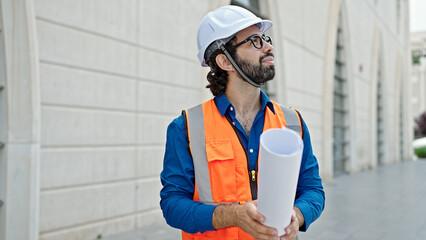 Young hispanic man architect holding blueprints looking around at construction place