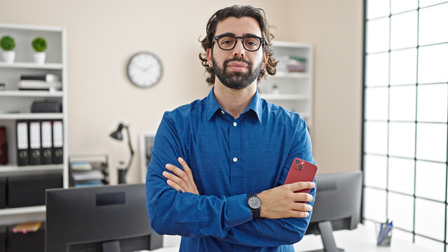 Young hispanic man business worker standing with arms crossed gesture holding smartphone at the office