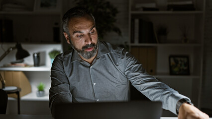 Mature hispanic man with grey beard working late in a dark office interior, focusing intently on his laptop screen.