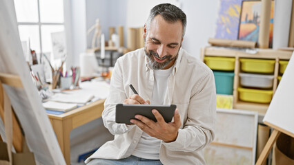 Mature hispanic man with grey beard smiling as he draws on a tablet in a bright art studio.