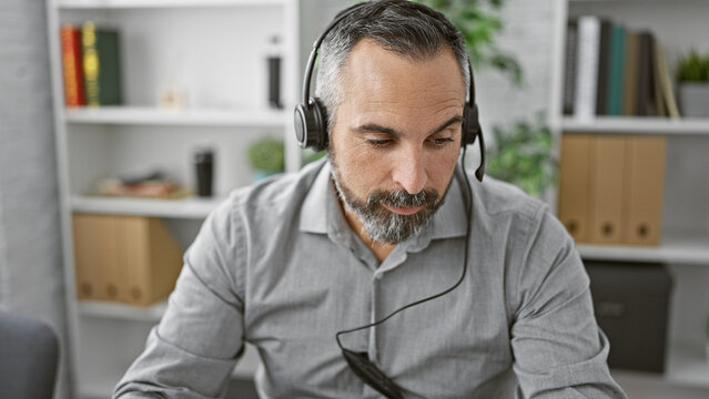 A mature hispanic man with a beard working in an office, wearing headphones and looking down.