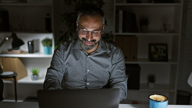 Handsome senior hispanic man with grey beard smiling at laptop in dark office room at night.