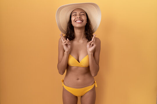 Young hispanic woman wearing bikini and summer hat gesturing finger crossed smiling with hope and eyes closed. luck and superstitious concept.