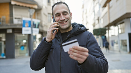 Smiling hispanic man using smartphone and holding credit card on urban city street