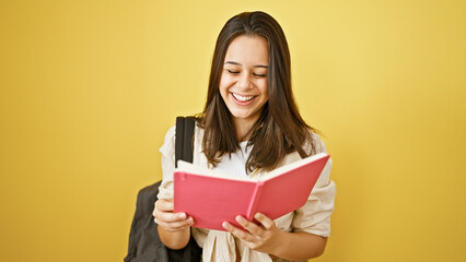 Joyful young hispanic woman with beautiful hairstyle standing confident, cheery, looking cool. casually dressed college student with backpack reading book. isolated on yellow wall background
