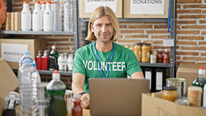 A blond man volunteers at a food donation center, sorting items indoors with a laptop.