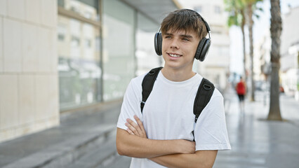 Caucasian teenage boy with headphones smiling outdoors on a sunny urban street while carrying a...
