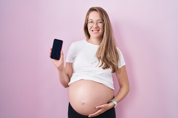 Young pregnant woman showing belly holding smartphone winking looking at the camera with sexy...