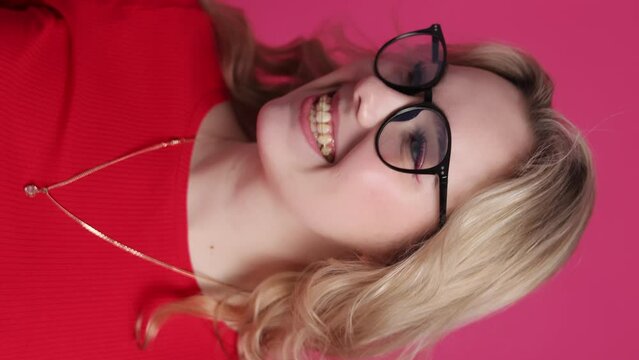 Cheerful young caucasian woman with transparent ceramic braces on teeth isolated on pink backdrop. Blond girl in eyeglasses looks at camera. Beauty, health and fashion concept. vertical video.