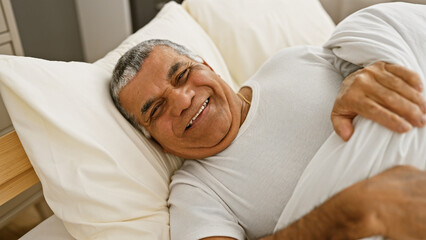 Obraz na płótnie Canvas A smiling middle-aged man with grey hair relaxes in bed, portraying comfort and happiness in a home bedroom setting.