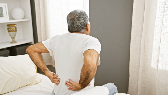 A middle-aged man with grey hair is experiencing back pain in his bedroom, depicting a common adult health issue.