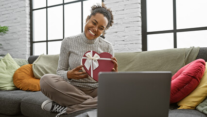 Smiling woman holding heart-shaped gift box while sitting on a couch indoors with laptop
